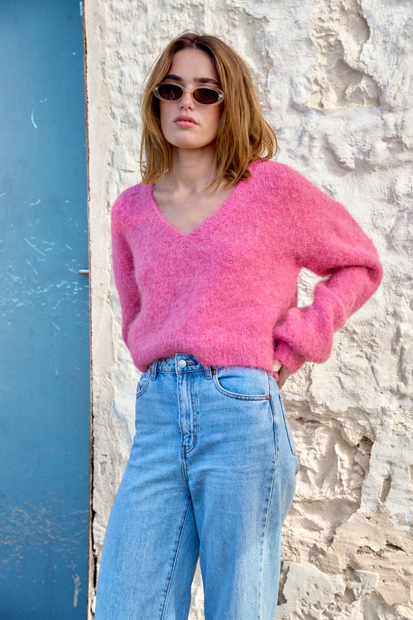 Woman wearing a pink sweater and blue jeans standing against a textured wall.