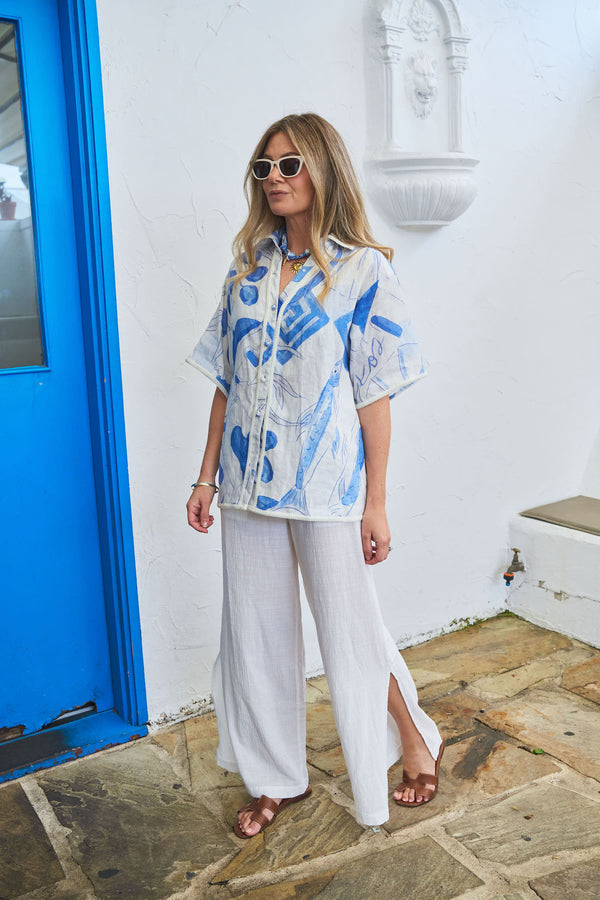 Jess Dempsey wearing a blue and white abstract patterned shirt and white pants standing in front of a blue door.